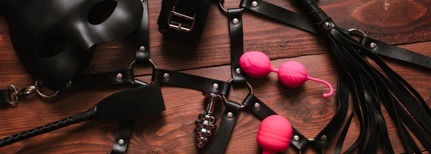 BDSM-Zubehör, schwarze Maske, Lederfesseln, Peitsche und rosa Liebeskugeln auf Holztisch