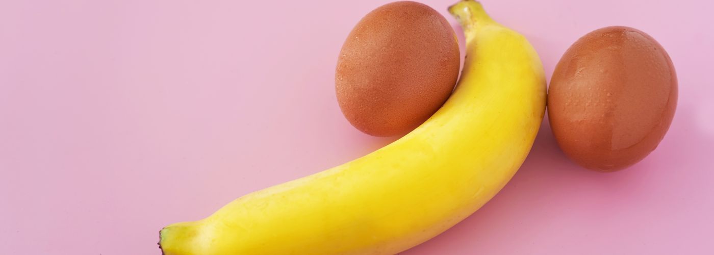Banane und zwei Eier symbolisch arrangiert auf rosa Hintergrund, sexual Wellness Bild
