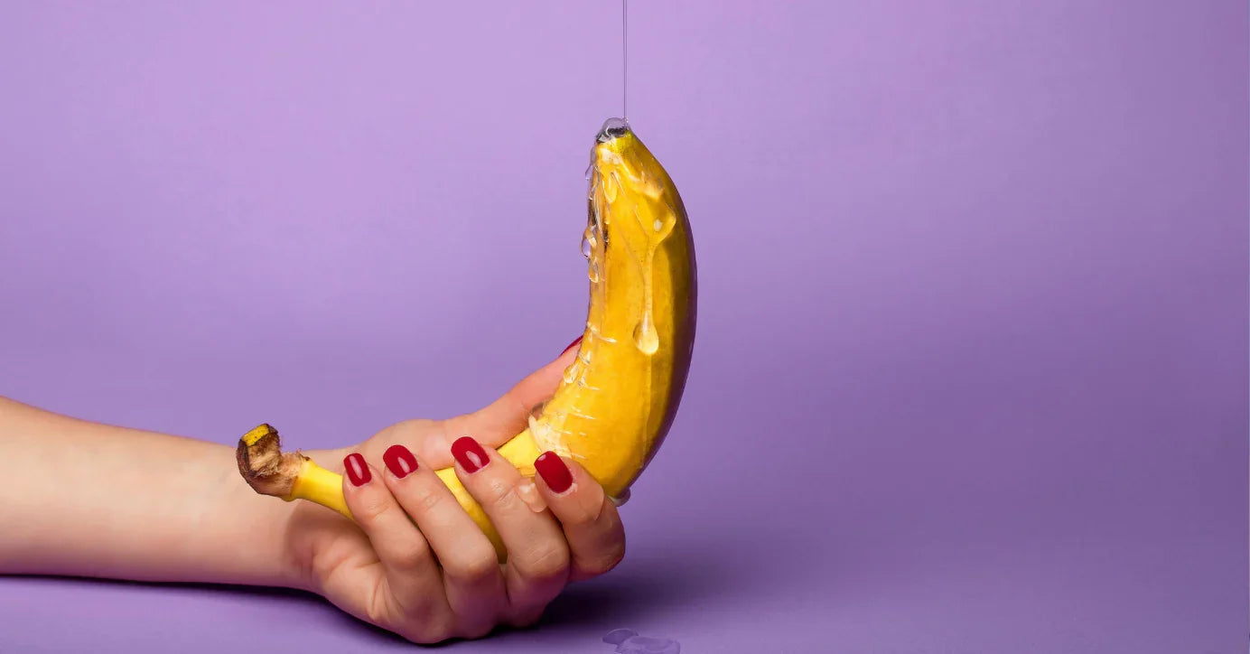 Hand hält Banane mit Gleitgel vor lila Hintergrund, Erotik, Sexspielzeug-Symbolik
