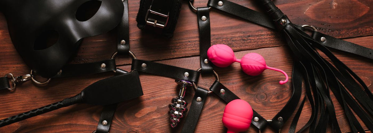 BDSM-Zubehör, schwarze Maske, Lederfesseln, Peitsche und rosa Liebeskugeln auf Holztisch