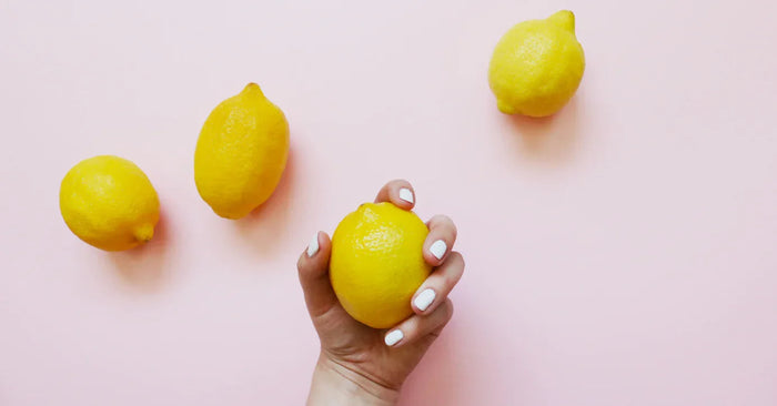 Hand hält Zitrone, drei Zitronen liegen auf rosa Hintergrund, Symbolbild Sexualität