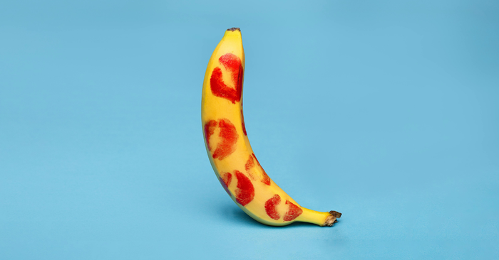 Banane mit roten Lippenstift-Kussabdrücken auf blauem Hintergrund, Symbol für Erotik und Liebesspiel