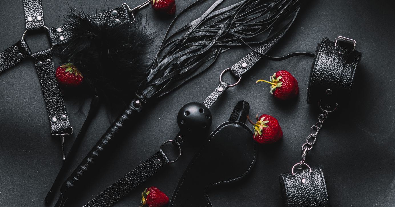 Schwarzes Bondage Set mit Handschellen, Augenmaske, Peitsche und Erdbeeren auf dunklem Hintergrund