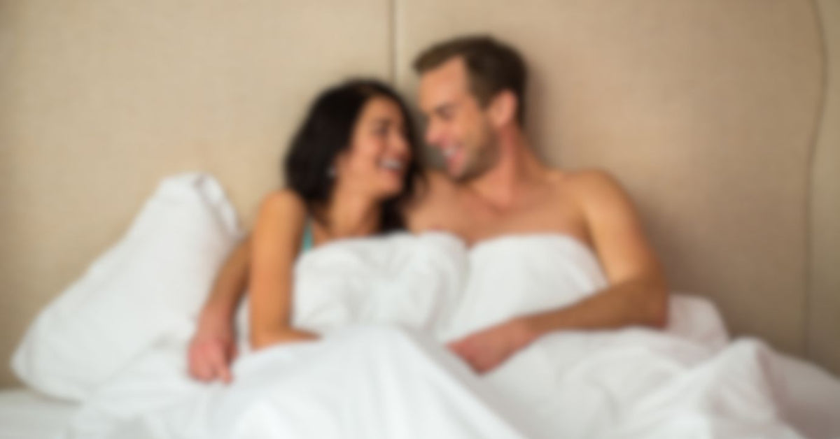 Lachendes Paar entspannt intim im Bett, weiße Bettwäsche, Symbole für Zweisamkeit und Liebesleben