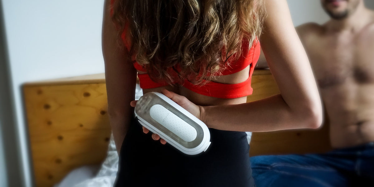 Frau hält Penisvibrator im Schlafzimmer, Mann sitzt im Hintergrund, erotische Stimmung