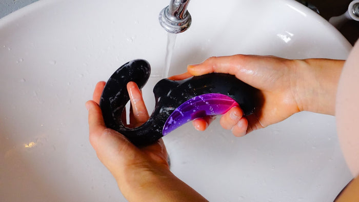 Person reinigt schwarzen und lila Vibrator unter fließendem Wasser am Waschbecken