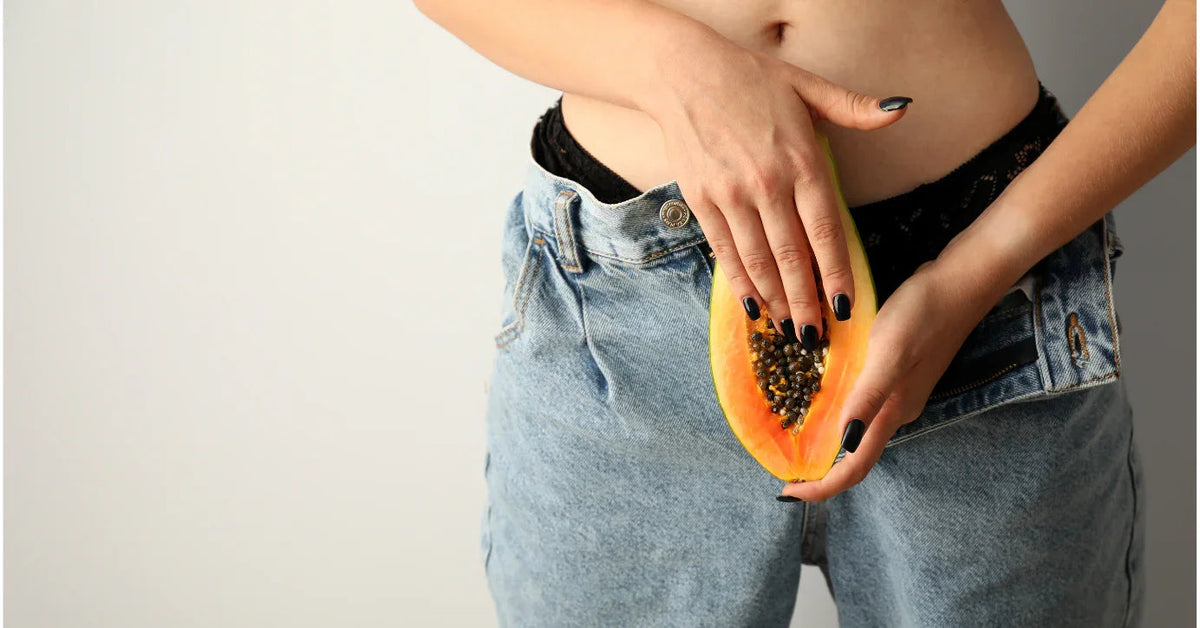 Frau mit aufgeknöpfter Jeans hält Papaya im Schritt, Sinnbild für Sexualität und Intimbereich