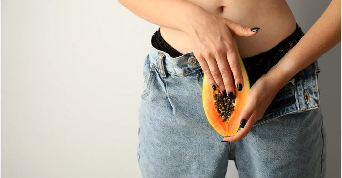Frau mit aufgeknöpfter Jeans hält Papaya im Schritt, Sinnbild für Sexualität und Intimbereich