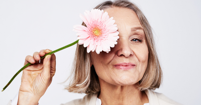 Ältere Frau hält rosa Blume vor das Auge, Symbol für Lebensfreude und Selbstbewusstsein