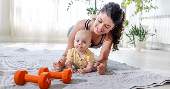 Mutter macht Fitness auf Matte mit Baby, zwei orange Hanteln im Vordergrund, helle Wohnung