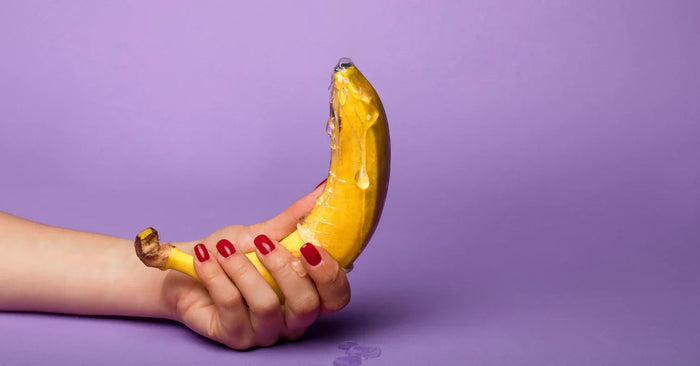 Hand mit roten Nägeln hält Banane mit Gleitgel vor lila Hintergrund, Symbol für Erotikartikel