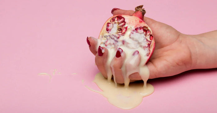 Frau mit lackierten Fingern hält halbierten Granatapfel mit cremigem Gleitgel auf rosa Hintergrund