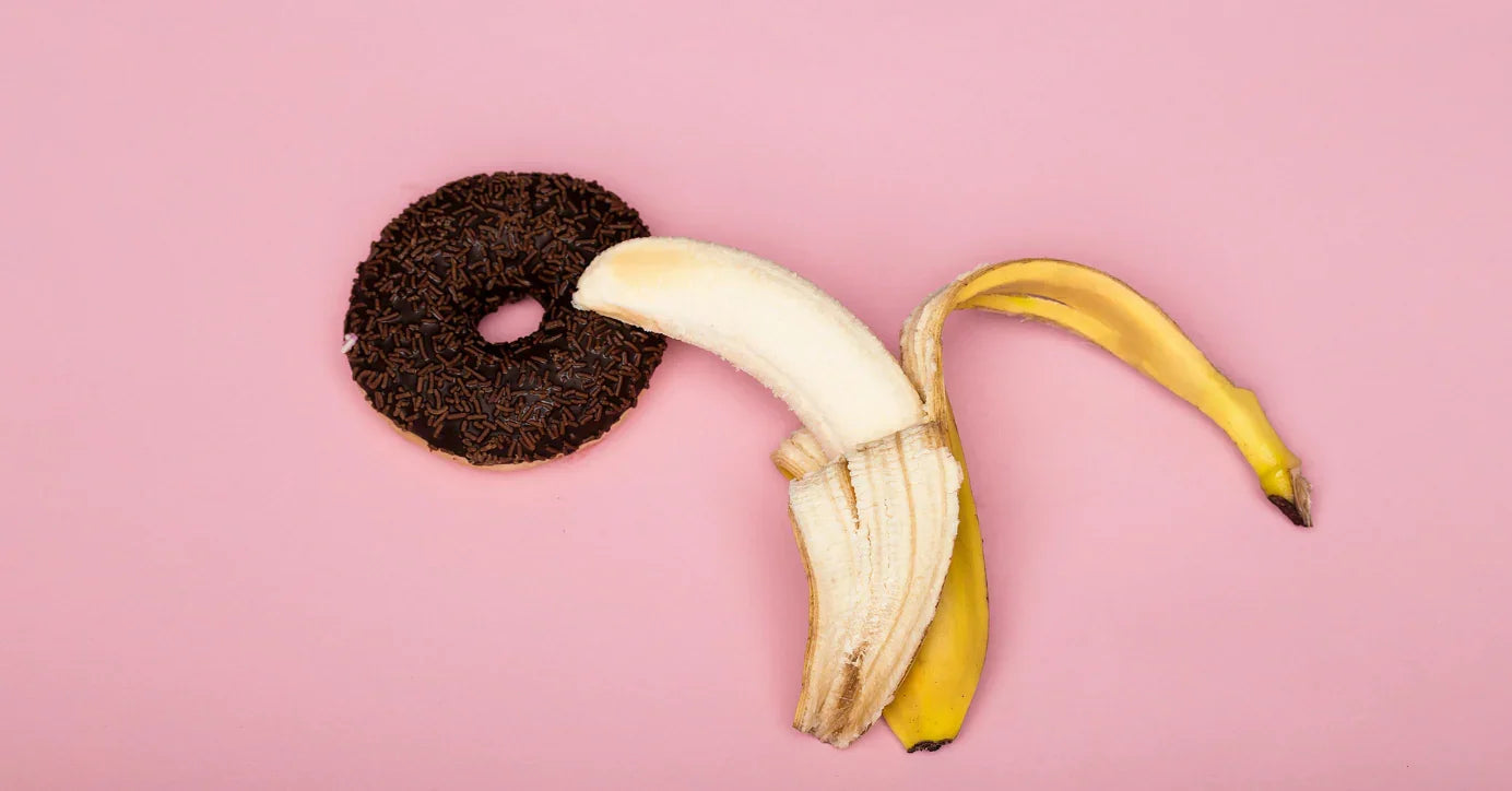 Banane und Schokodonut auf rosa Hintergrund, symbolisch für Erotik, Sexualität und Liebesleben