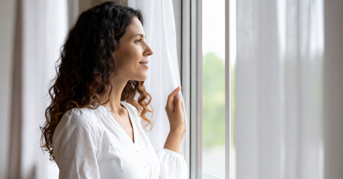 Frau mit lockigen Haaren blickt entspannt aus dem Fenster, sinnliche Stimmung, helle Gardinen